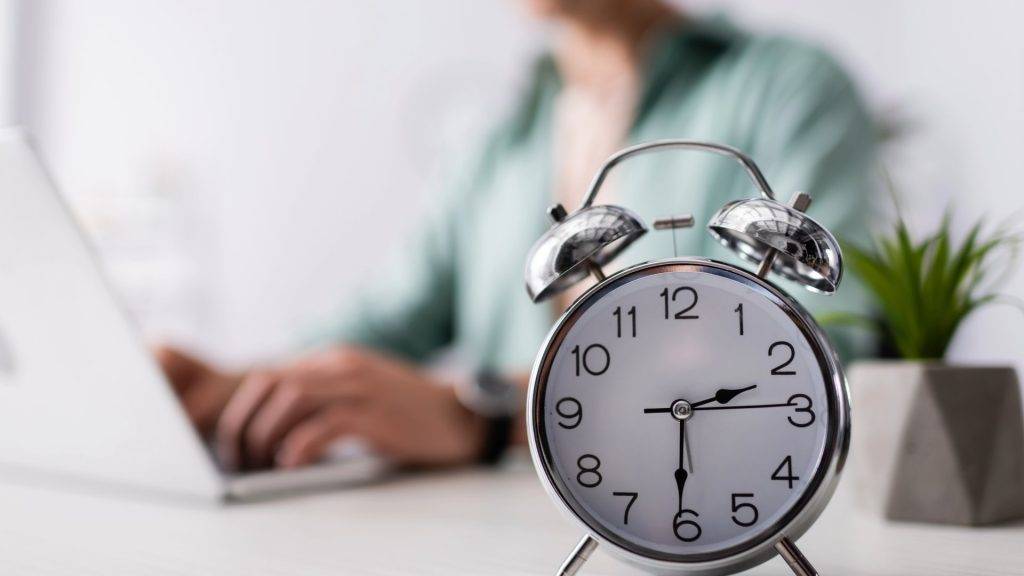 Silver alarm clock on a desk with a person working in the background, representing waiting time for an injection to work.