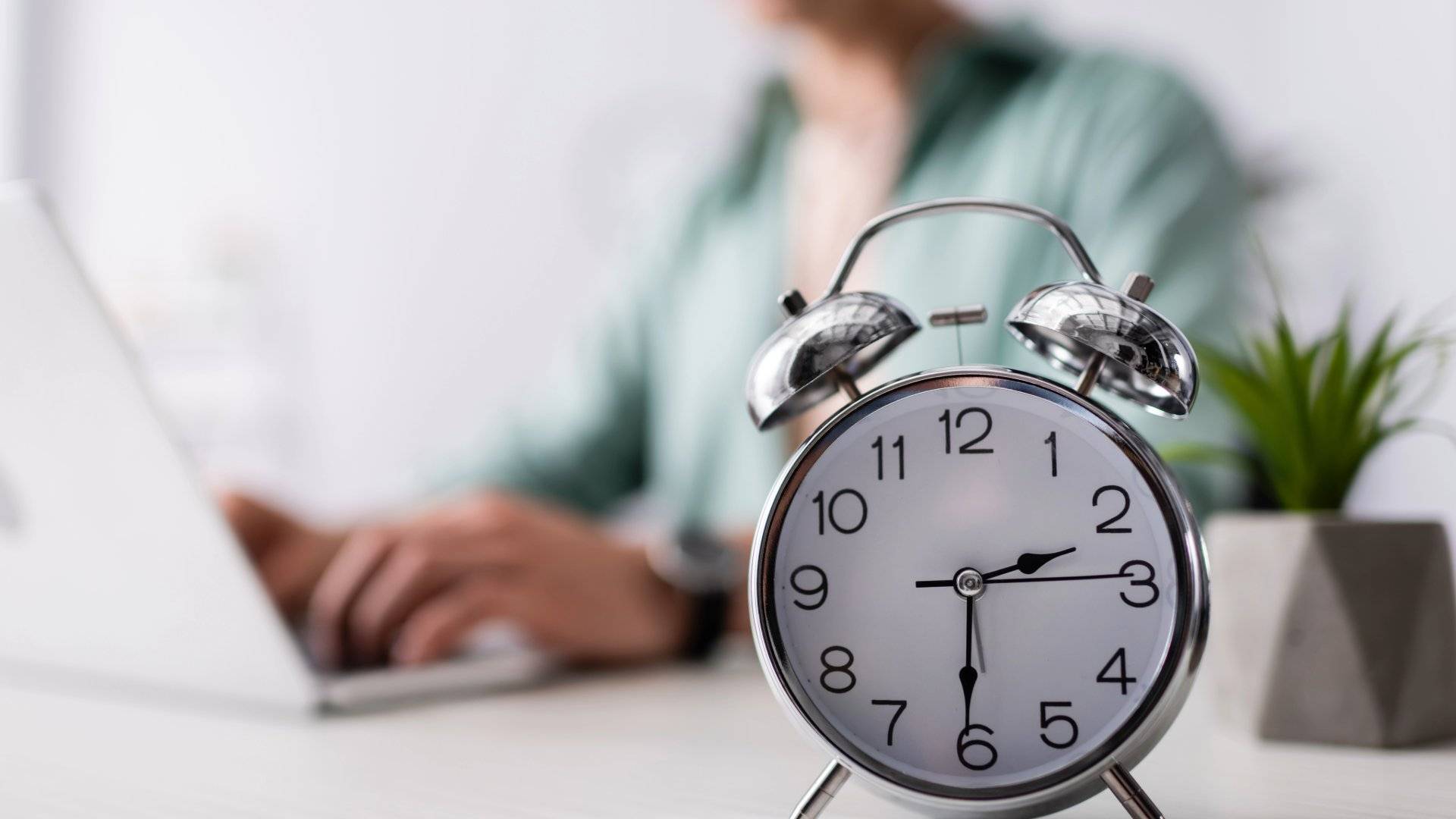 Silver alarm clock on a desk with a person working in the background, representing waiting time for an injection to work.