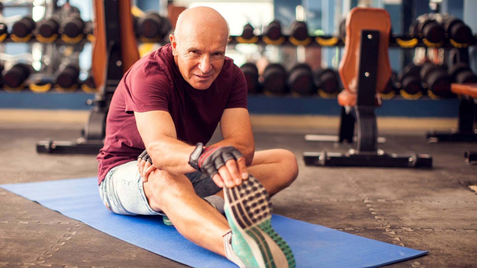 Older man stretching on a blue exercise mat in a gym with weights and benches in the background